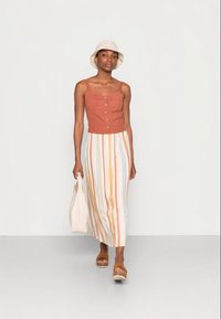 Top cropped de cor ferrugem com botões e uma saia longa listrada em branco, amarelo e laranja. A modelo leva uma bolsa branca e usa sandálias castanhas.
