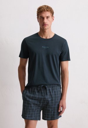 Man wearing dark blue short-sleeve T-shirt with small logo and navy blue checkered shorts with drawstring, standing against a plain white wall.