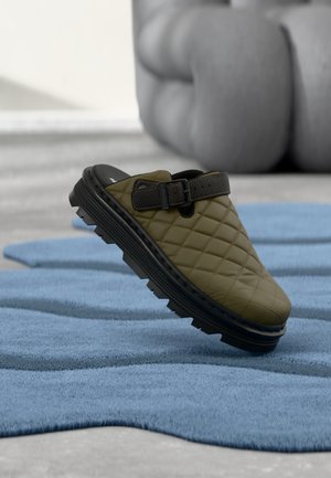 Sabot vert olive matelassé à enfiler avec une semelle épaisse noire et une sangle ajustable, posé sur un tapis bleu texturé dans un décor intérieur moderne.