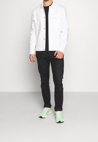 Muž ve bílém button-up saku, černém tričku, černých kalhotách a světlých zelených teniskách stojící na pozadí s hladkou bílou barvou.