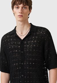 Camisa de encaje negra con cuello abotonado, que presenta un patrón intrincado y mangas cortas. Ligera y semi-translúcida con botones negros.