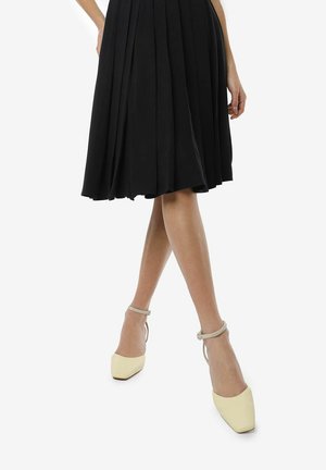 Vestido midi negro con pliegues y un diseño fluido, llevado con tacones de punta amarillos pálidos que presentan una correa en el tobillo plateada.
