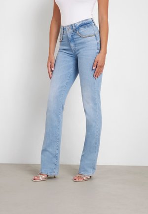 Vrouw met lichtblauwe jeans met hoge taille en steentjes op de zakken, gecombineerd met een wit shirt en zilveren sandalen met bandjes.
