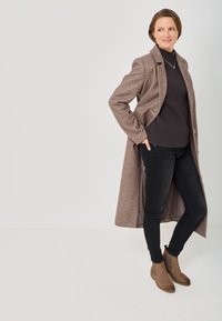 Lungo cappotto di lana marrone con colletto a punta sopra un maglione nero, abbinato a jeans skinny neri e stivaletti marrone.