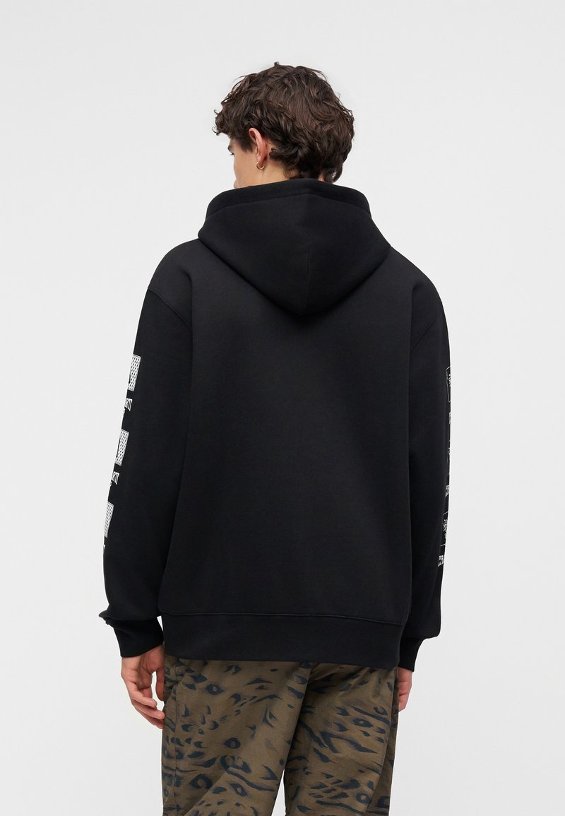 Junge Person mit lockigem dunklem Haar, die einen schwarzen Hoodie mit bedruckten Ärmeln und braune Tarnhosen trägt, von hinten auf einfarbigem Hintergrund.