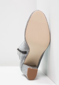 Zapato de tacón plateado y brillante con un upper texturizado, cremallera lateral y suela de goma beige. Punta ligeramente redondeada y forma afilada.