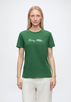 Femme aux cheveux blonds raides portant un t-shirt vert Tommy Hilfiger et un pantalon blanc, debout devant un fond clair uni.