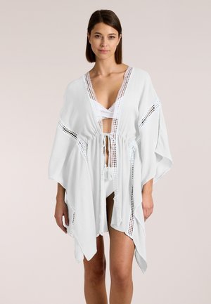 Frau trägt ein weißes offen vorne getragenes Kaftan mit Häkelbesatz, in der Taille gebunden, über weißer Badebekleidung vor hellem, einfarbigem Hintergrund.