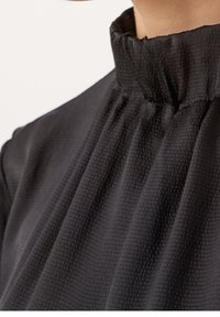 Czarna teksturowana bluzka z wysokim, falbankowym kołnierzem i miękkim drapowaniem. Na tkaninie delikatny wzór podkreślający jej lekki charakter.
