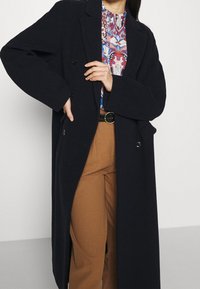 Cappotto lungo in misto lana blu navy con una vestibilità snella e design doppiopetto, dettagli con bottoni. Indossato sopra una camicia a stampa colorata e pantaloni marroni.