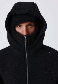 Personne portant une veste à capuche noire zippée jusqu'à couvrir la bouche et le nez, ne montrant que les yeux et le front sur un fond uni.