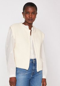 Vrouw in een textuurachtige crèmekleurige mouwloze sweater vest over een witte blouse en blauwe jeans, met gouden hangoorbellen.