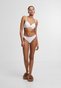 Witte bh met kantafwerking en bijpassende bikini briefs, met een gladde textuur. Model draagt bruin harig slippers. Eenvoudige achtergrond.