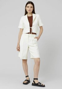 Veste blanche raccourcie à manches courtes et à lien devant, associée à un short blanc à longueur de genoux et des sandales noires à lanières. Tissu lisse et léger.