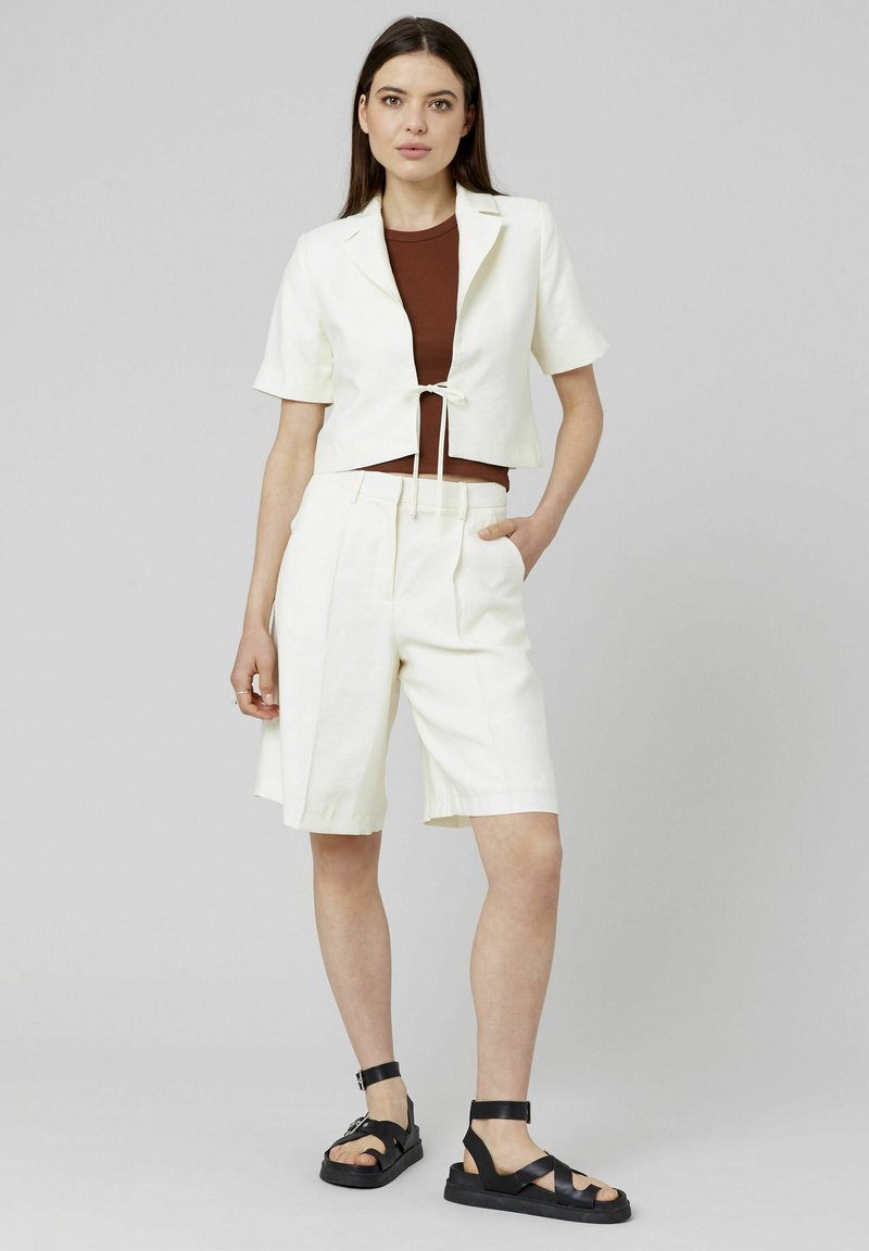 Veste blanche raccourcie à manches courtes et à lien devant, associée à un short blanc à longueur de genoux et des sandales noires à lanières. Tissu lisse et léger.