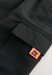 Schwarzer Stoff mit glatter Textur, ausgestattet mit einem umgeschlagenen Bund und einem orangefarbenen rechteckigen Etikett mit schwarzer Logodetails.