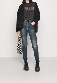 Cardigan de punto negro sobre una camiseta gráfica gris con el texto "VACATION", combinado con jeans ajustados azules, botas negras con cordones y un bolso de cuadros.