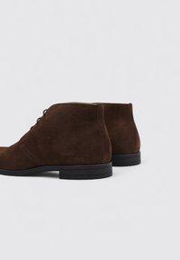 Botas de tobillo de ante marrón con una textura suave, punta redonda y suela de goma negra duradera. Presenta un detalle de cordones minimalista en la parte frontal.
