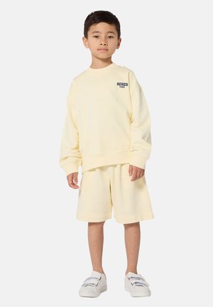 Gelber Pullover mit Rundhalsausschnitt und lockerem Schnitt, kombiniert mit passenden gelben Shorts. Weiße Sneakers mit Klettverschlüssen.