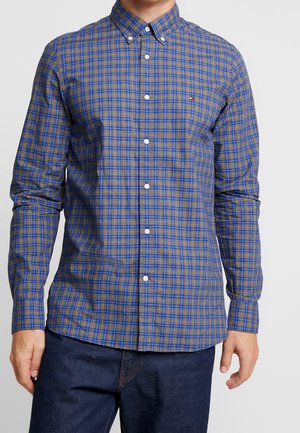 Mann trägt blau-gelbes kariertes Hemd mit langen Ärmeln und dunkelblaue Jeans vor einem einfarbigen Hintergrund.