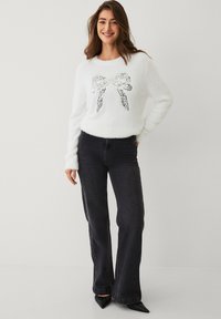 Witte, pluizige trui met een strik van pailletten op de voorkant, gecombineerd met donkere uitlopende spijkerbroeken en zwarte schoenen met een puntige neus.