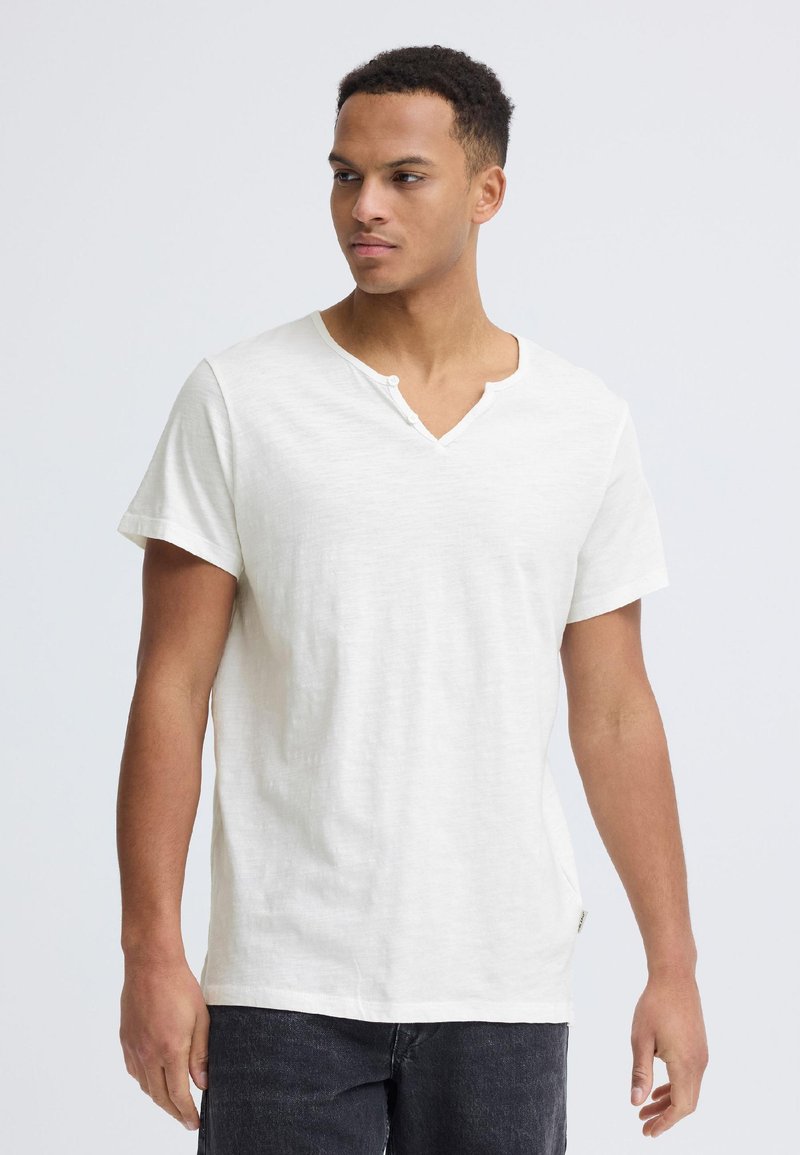 Junger Mann trägt ein schlichtes weißes Henley-Shirt mit kurzen Ärmeln und dunkle Jeans, steht vor einem schlichten hellgrauen Hintergrund.