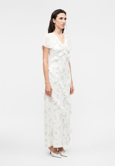 Γυναίκα που φοράει λευκό μακρύ φόρεμα με λουλούδια, λεπτομέρειες με βολάν και κοντά φουντωτά μανίκια, συνδυασμένο με λευκά μυτερά ψηλοτάκουνα παπούτσια.