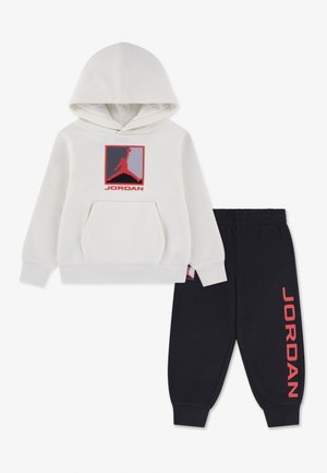Witte hoodie met een rood-zwart Jordan-logo en zwarte broek met rode Jordan-letters langs het been.