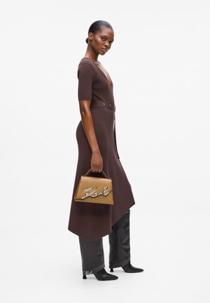 Woman in brown belted dress and black boots holding a tan handbag with silver signature detail, standing against a white background.