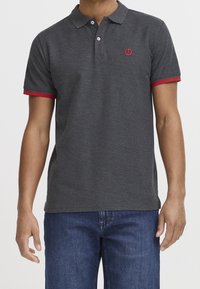 Muž v tmavě šedém polo tričku s červeným lemem na rukávech a malým červeným logem, v kombinaci s modrými džíny.