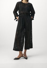 Mustasyhtö, joka koostuu läpinäkyvästä, kuviollisesta pitkähihasta yläosasta ja leveistä housuista. Asusteena teräväkärkiset mustat korkokengät, joissa on hopeinen yksityiskohta.