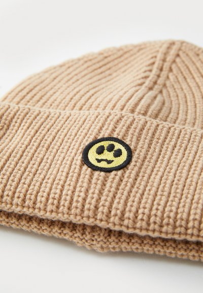 Berretto beige lavorato a maglia con texture a coste, dotato di un cerchio nero con un design di faccina sorridente gialla.