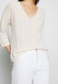 Kremowy sweter dzianinowy z dekoltem w kształcie litery V i rękawami o długości trzech czwartych, cechujący się luźnym krojem i teksturowanym wzorem, zestawiony z białymi dżinsami.