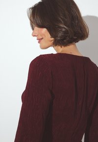 Abito borgogna strutturato con pieghe verticali e maniche lunghe. Il colletto è rotondo e il tessuto ha una leggera lucentezza.