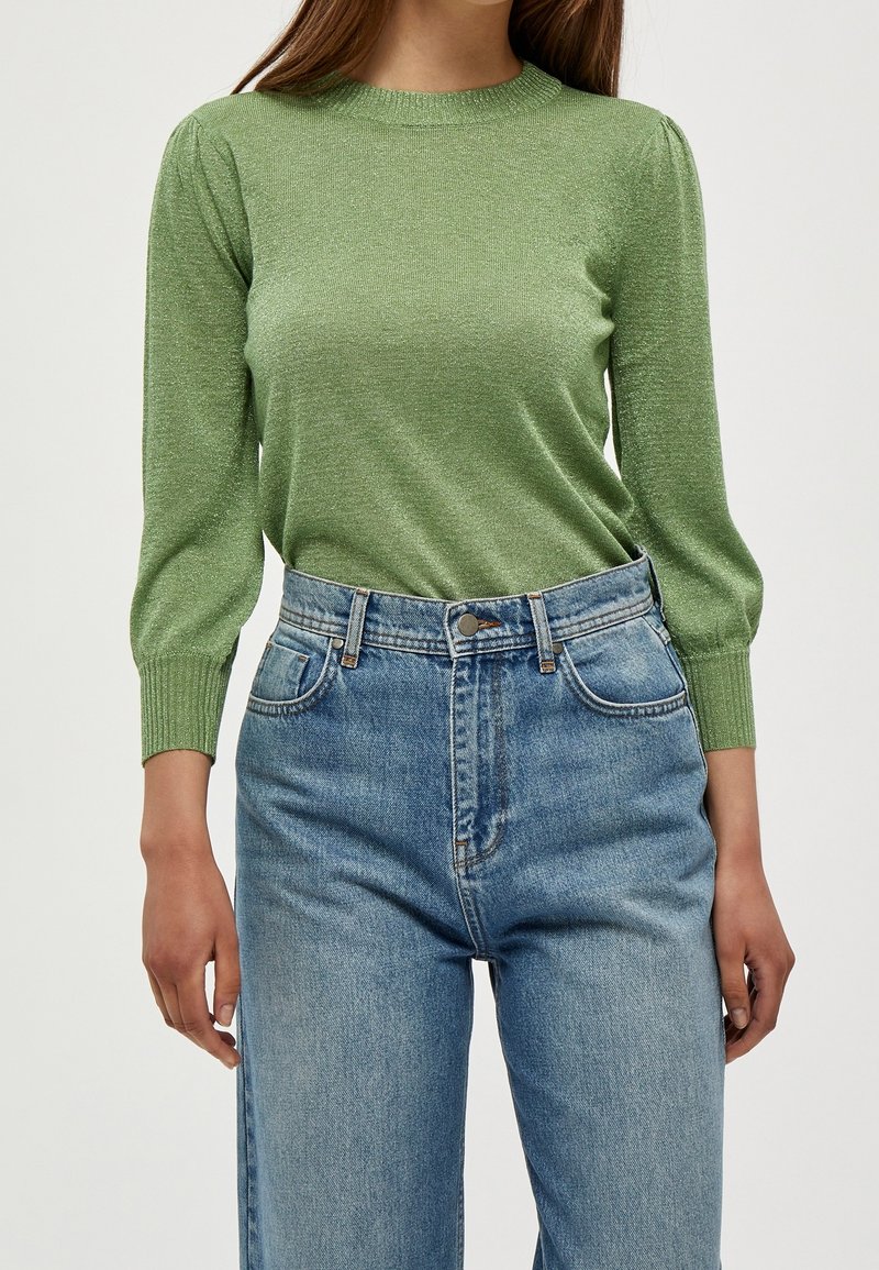 Pull en maille métallique verte avec col et poignets côtelés, associé à un jean en denim bleu clair taille haute avec des poches classiques.