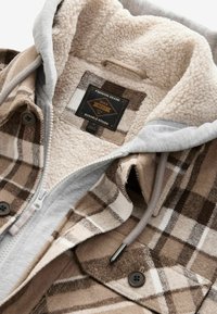 Veste à carreaux marron et beige avec doublure en polaire, capuche grise avec cordons, poches à boutons et une étiquette noire à l’intérieur du col.