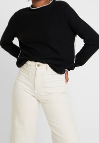 Svart stickad tröja med vit kant vid kragen och ärmsluten, kombinerad med högmidjade krämfärgade vida jeans med framfickor.