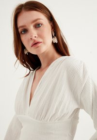 Abito bianco con texture, scollatura a V profonda e vita arricciata. Modella che indossa orecchini trasparenti a forma di foglia. Sfondo semplice e di colore chiaro.