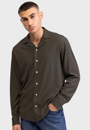 Νεαρός με κοντά μαλλιά, φορούν σκούρο ελιά πουκάμισο με κουμπιά και μπλε τζιν, στέκεται με το ένα χέρι στην τσέπη του μπροστά από ένα απλό φόντο.