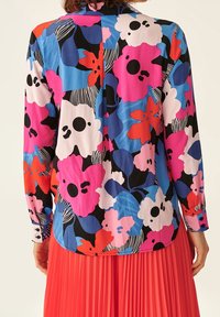 Donna che indossa una blusa floreale colorata con fiori rosa, blu, rossi e bianchi abbinata a una vivace gonna plissettata rossa.
