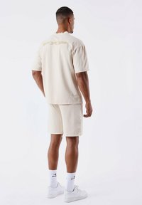 Set di t-shirt e pantaloni corti in cotone beige. La t-shirt presenta un discreto logo "PXP" sul retro, abbinata a sneaker bianche e calzini a righe.