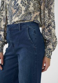 Temno modre kavbojke iz jeansa z teksturirano površino, visokim pasom, sprednjimi žepi in ravnimi hlačnicami. Kombinirane s potiskano bluzo.