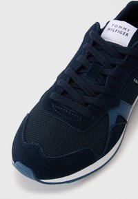 Sneaker bleumarin cu un material de plasă texturat, accente din piele întoarsă și șireturi plate. Include o etichetă albă cu logo pe limbă și o talpă din cauciuc.