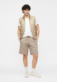 Colete acolchoado bege com gola alta, t-shirt branca, calções bege estampados e ténis creme. Modelo posa sobre fundo neutro.