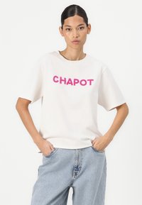 Balta trumpomis rankovėmis marškinėliai su ryškiu rožiniu "CHAPOT" užrašu, pagaminti iš minkštos medžiagos, laisvės stiliaus, derinami su šviesiai mėlynomis džinsomis.