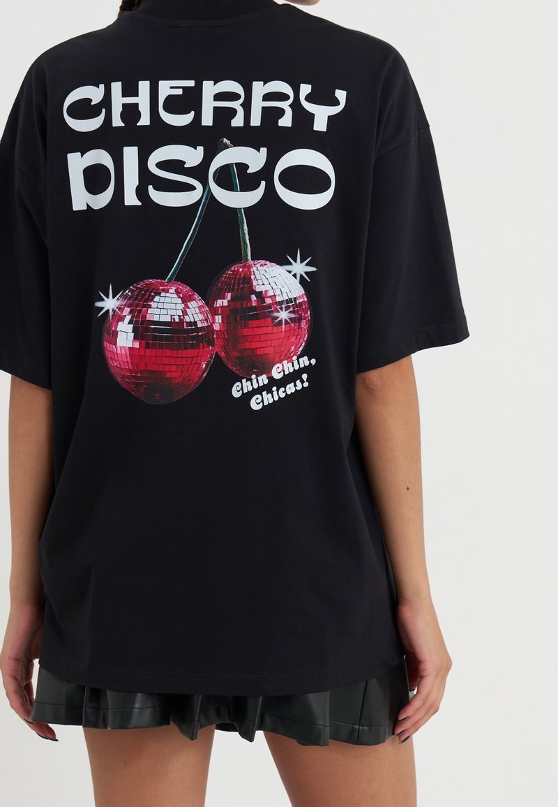 T-shirt noir oversize avec le texte "CHERRY DISCO" et deux cerises boule à facettes, porté par une personne avec une jupe noire visible en dessous.