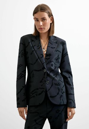Donna con blazer nero su misura e pantaloni con testo grafico nero sparso e simboli, in piedi su sfondo bianco.