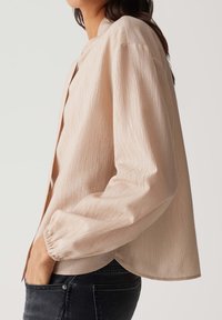 Beige long-sleeve shirt with a textured, pleated design, button closure, and relaxed fit. Features gathered cuffs and a curved hem.