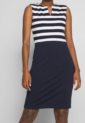 Vrouw draagt een mouwloze marineblauwe jurk met witte horizontale strepen op de bovenste helft en een effen marineblauwe onderkant tegen een grijze achtergrond.