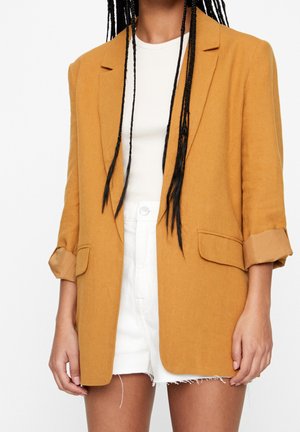 Person wearing a mustard yellow blazer with rolled sleeves, white ribbed top, and white high-waisted shorts with frayed hems.